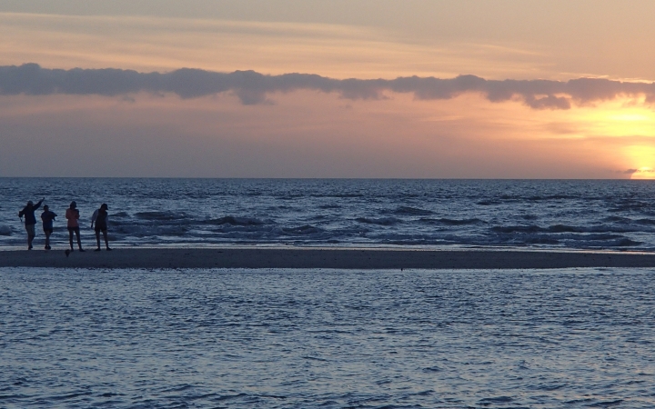 Four people stand on a small peninsula surrounded by blue water. In the background, the sun is setting on the horizon, illuminating the sky in soft shades in yellow, orange, pink and purple. 